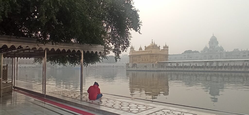 The Golden Temple (Harmandir Sahib) surrounded by the sacred Amrit Sarovar lake, its glistening gold-plated walls and dome reflecting in the water. Devotees walk along the marble walkway, and the temple’s intricate architecture is visible under a clear blue sky.
